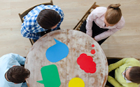 Overhead picture looking down at four people sitting around a table with a green paint splash, a red paint splash a blue paint splash and a yellow paint splash.